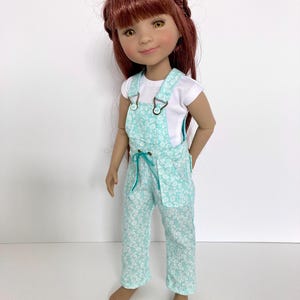 May include: A doll with long auburn hair and brown eyes wearing a white short-sleeved shirt under light teal overalls. The overalls feature a white floral pattern, metal buckles on the straps, and a drawstring waist with teal cord accents.