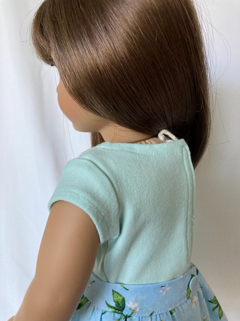 Puede incluir: Una mu&ntilde;eca con cabello casta&ntilde;o largo, que lleva una camiseta de manga corta azul claro y una falda floral azul. La falda tiene un estampado floral blanco. La mu&ntilde;eca est&aacute; mirando hacia la izquierda, con el lado de su cara visible.