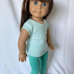 Puede incluir: Mu&ntilde;eca con cabello casta&ntilde;o y ojos azules, vestida con una camiseta azul claro, leggings turquesas y sandalias rosas con lunares blancos. La mu&ntilde;eca est&aacute; de pie frente a un fondo blanco.
