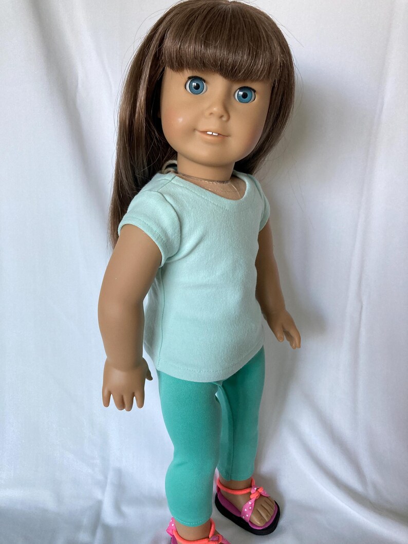 Puede incluir: Mu&ntilde;eca con cabello casta&ntilde;o y ojos azules, vestida con una camiseta azul claro, leggings turquesas y sandalias rosas con lunares blancos. La mu&ntilde;eca est&aacute; de pie frente a un fondo blanco.