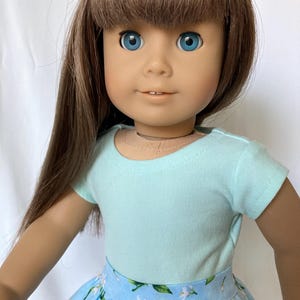 Puede incluir: Una mu&ntilde;eca con cabello casta&ntilde;o y ojos azules, vestida con una camiseta azul claro y una falda floral azul. La mu&ntilde;eca tiene una tez clara y mira directamente al espectador. El cabello de la mu&ntilde;eca est&aacute; peinado con flequillo.