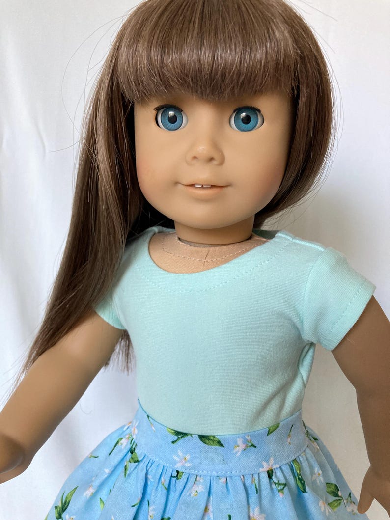 Puede incluir: Una mu&ntilde;eca con cabello casta&ntilde;o y ojos azules, vestida con una camiseta azul claro y una falda floral azul. La mu&ntilde;eca tiene una tez clara y mira directamente al espectador. El cabello de la mu&ntilde;eca est&aacute; peinado con flequillo.