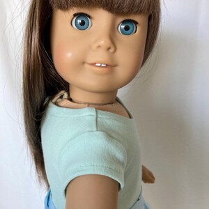 Puede incluir: Una mu&ntilde;eca con cabello casta&ntilde;o y ojos azules, vestida con una camiseta azul claro y una falda con estampado floral azul. La mu&ntilde;eca tiene una expresi&oacute;n sonriente y mira hacia el espectador.