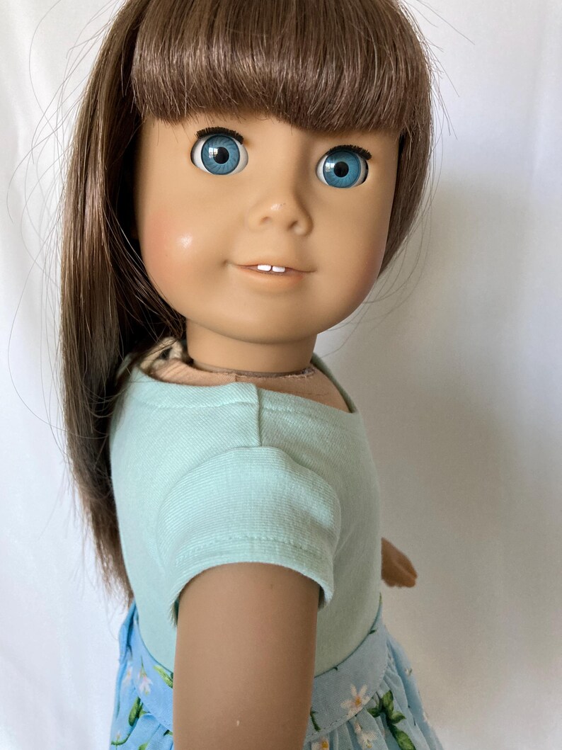 Puede incluir: Una mu&ntilde;eca con cabello casta&ntilde;o y ojos azules, vestida con una camiseta azul claro y una falda con estampado floral azul. La mu&ntilde;eca tiene una expresi&oacute;n sonriente y mira hacia el espectador.
