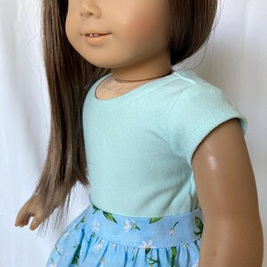 Puede incluir: Una mu&ntilde;eca vestida con una camiseta azul claro y una falda azul con un estampado floral blanco. La mu&ntilde;eca tiene pelo largo casta&ntilde;o y ojos azules. El conjunto est&aacute; dise&ntilde;ado para una mu&ntilde;eca.