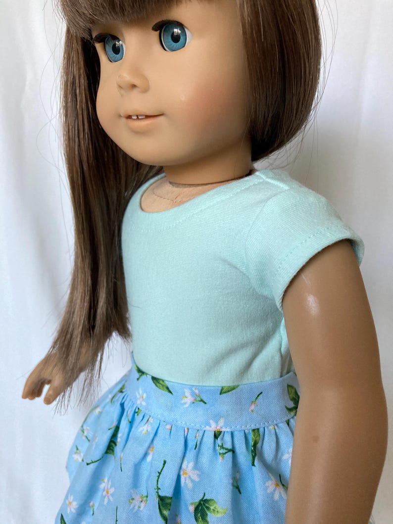 Puede incluir: Una mu&ntilde;eca vestida con una camiseta azul claro y una falda azul con un estampado floral blanco. La mu&ntilde;eca tiene pelo largo casta&ntilde;o y ojos azules. El conjunto est&aacute; dise&ntilde;ado para una mu&ntilde;eca.