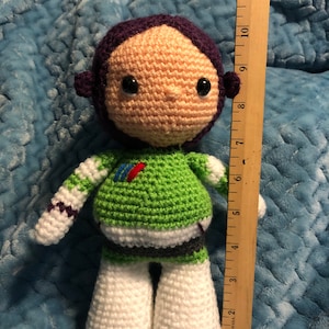 K&ouml;nnte beinhalten: Handgefertigte H&auml;kelpuppe, die Buzz Lightyear darstellt. Die Puppe ist etwa 25 cm gro&szlig; und hat einen gr&uuml;n-wei&szlig;en Raumanzug, einen lila Helm und ein beiges Gesicht mit schwarzen Augen. Die Puppe ist aus Garn gefertigt.