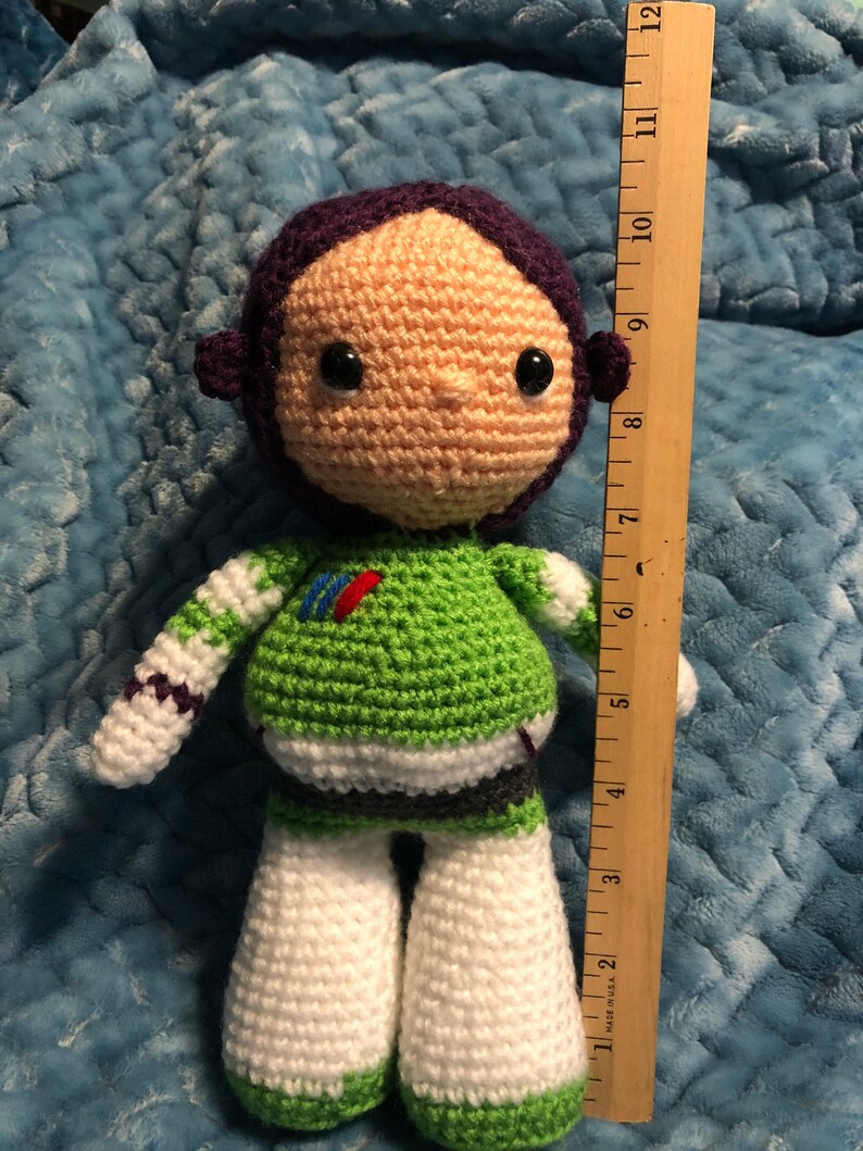 K&ouml;nnte beinhalten: Handgefertigte H&auml;kelpuppe, die Buzz Lightyear darstellt. Die Puppe ist etwa 25 cm gro&szlig; und hat einen gr&uuml;n-wei&szlig;en Raumanzug, einen lila Helm und ein beiges Gesicht mit schwarzen Augen. Die Puppe ist aus Garn gefertigt.