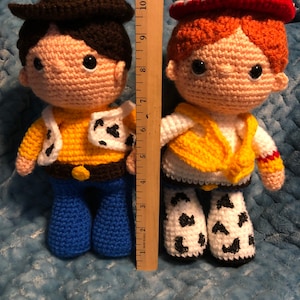 K&ouml;nnte beinhalten: Zwei handgefertigte H&auml;kelpuppen, eine, die einem Cowboy mit braunem Hut, gelbem Hemd und blauer Jeans &auml;hnelt, und die andere einem Cowgirl mit rotem Hut, gelber Weste und Chaps mit Kuhmuster. Die Puppen sind etwa 25 cm gro&szlig;.