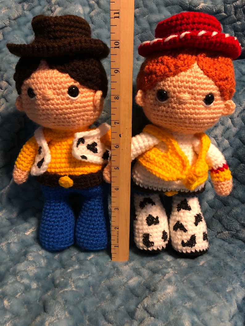 K&ouml;nnte beinhalten: Zwei handgefertigte H&auml;kelpuppen, eine, die einem Cowboy mit braunem Hut, gelbem Hemd und blauer Jeans &auml;hnelt, und die andere einem Cowgirl mit rotem Hut, gelber Weste und Chaps mit Kuhmuster. Die Puppen sind etwa 25 cm gro&szlig;.