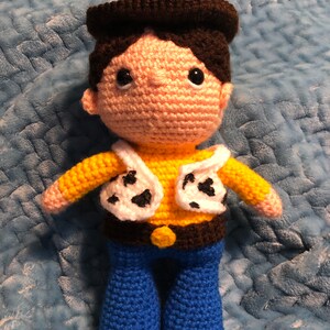 K&ouml;nnte beinhalten: Geh&auml;kelte Puppe, die einem Cowboy-Charakter &auml;hnelt. Die Puppe hat einen braunen Hut, Haare und einen G&uuml;rtel, ein gelbes Hemd, eine wei&szlig;e Weste mit schwarzen Flecken und eine blaue Hose. Die Puppe hat schwarze Augen und ein pfirsichfarbenes Gesicht und H&auml;nde.
