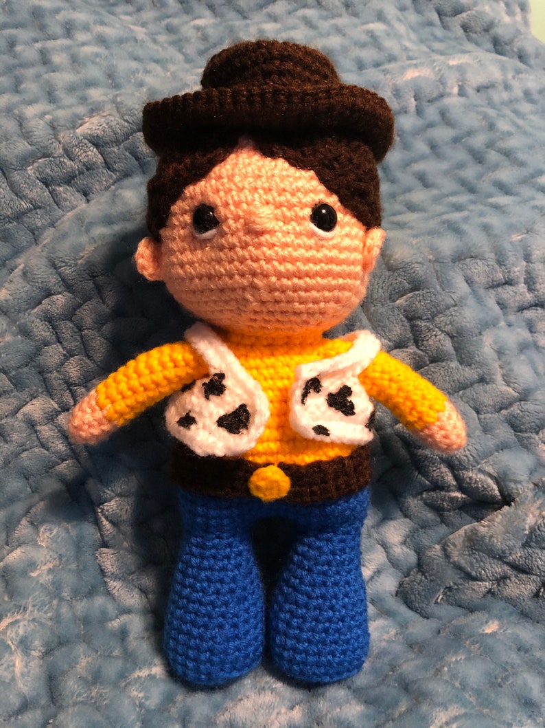 K&ouml;nnte beinhalten: Geh&auml;kelte Puppe, die einem Cowboy-Charakter &auml;hnelt. Die Puppe hat einen braunen Hut, Haare und einen G&uuml;rtel, ein gelbes Hemd, eine wei&szlig;e Weste mit schwarzen Flecken und eine blaue Hose. Die Puppe hat schwarze Augen und ein pfirsichfarbenes Gesicht und H&auml;nde.