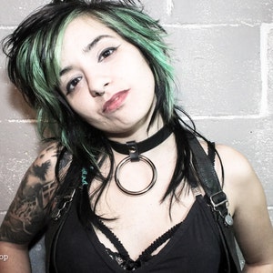 May include: A woman with green and black hair is wearing a black choker with a large silver ring. She is wearing a black tank top with straps and a black leather harness. She has a tattoo on her arm.