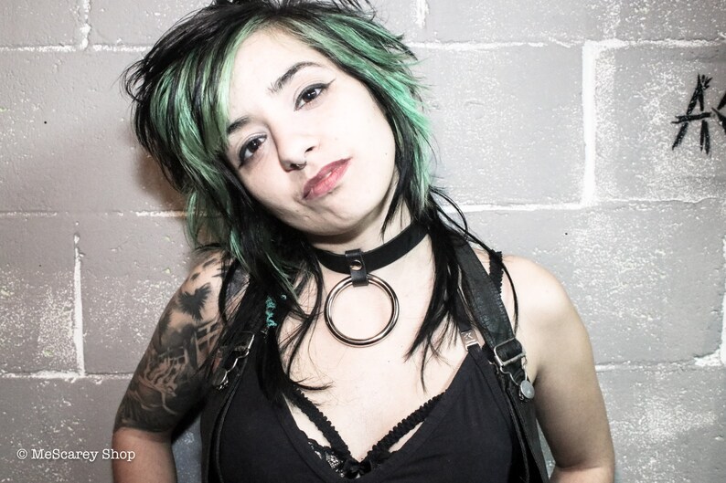 May include: A woman with green and black hair is wearing a black choker with a large silver ring. She is wearing a black tank top with straps and a black leather harness. She has a tattoo on her arm.