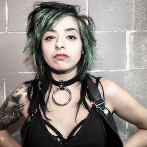 May include: A woman with green hair and a black choker with a large silver ring. She is wearing a black tank top and has a tattoo on her arm.