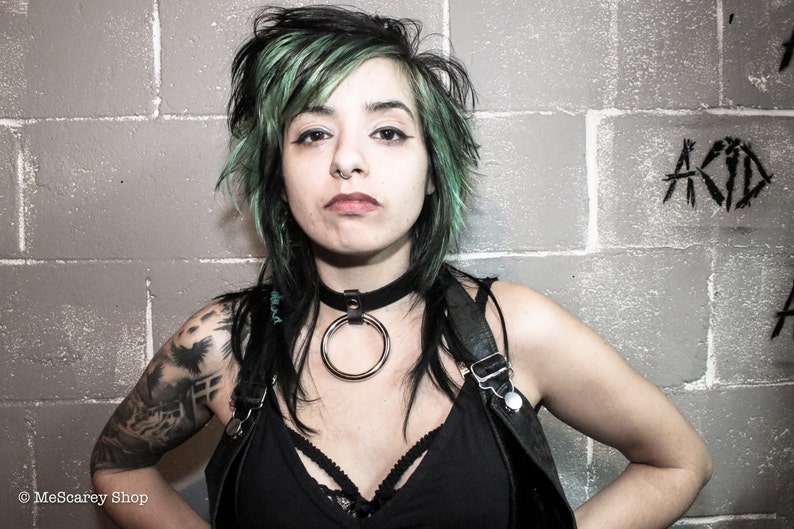 May include: A woman with green hair and a black choker with a large silver ring. She is wearing a black tank top and has a tattoo on her arm.