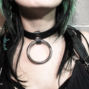 May include: A black leather choker with a silver ring attached. The choker is worn by a person with black and green hair.