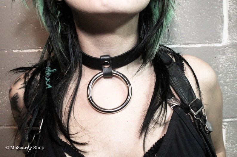 May include: A black leather choker with a silver ring attached. The choker is worn by a person with black and green hair.