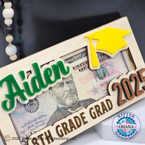 Puede incluir: Un marco de madera con un recorte de gorro de graduaci&oacute;n, un nombre verde "Aiden" y el texto "6th Grade Grad 2025". Un billete de d&oacute;lar es visible en el marco.