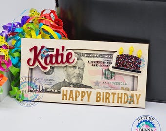 May include: A wooden birthday gift holder with the name "Katie" in red script. The holder contains a $50 bill and a small cake cutout. The words "HAPPY BIRTHDAY" are in gold lettering. Colorful ribbons are in the background.