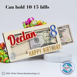 May include: Wooden money holder with the name "Declan" and the number "8" in blue. The holder has a slot for bills and says "Happy Birthday".