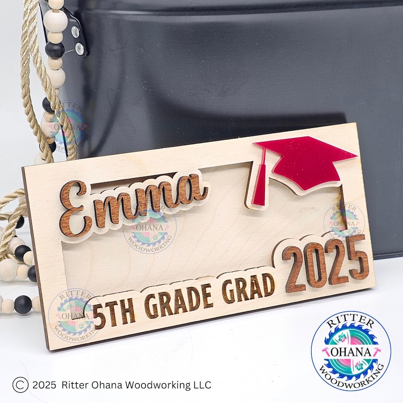 Puede incluir: Un letrero de madera con el nombre "Emma" y el texto "5th Grade Grad 2025". Un gorro de graduaci&oacute;n rojo est&aacute; en el letrero. El letrero est&aacute; hecho por Ritter Ohana Woodworking.