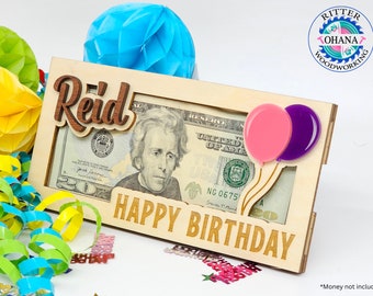 May include: Wooden money holder with a slot for a bill, featuring a pink and purple balloon design. The holder has a personalized name "Reid" and the words "Happy Birthday".
