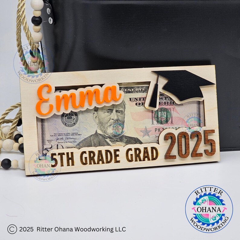 Puede incluir: Un letrero de madera con un recorte de gorro de graduaci&oacute;n, un billete de d&oacute;lar y el texto "Emma 5th Grade Grad 2025". El letrero est&aacute; hecho de madera clara con acentos naranja y marr&oacute;n.