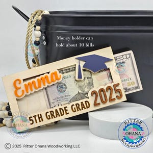 Puede incluir: Un portamonedas de madera con un dise&ntilde;o de gorro de graduaci&oacute;n. El portamonedas tiene una ranura para billetes y est&aacute; personalizado con el nombre "Emma" y el texto "5th Grade Grad 2025".
