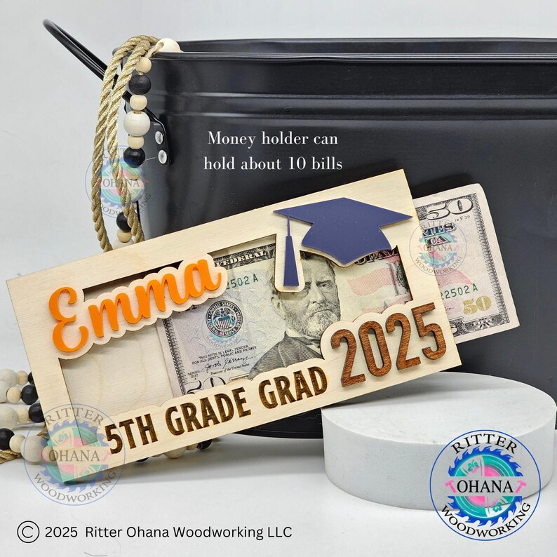 Puede incluir: Un portamonedas de madera con un dise&ntilde;o de gorro de graduaci&oacute;n. El portamonedas tiene una ranura para billetes y est&aacute; personalizado con el nombre "Emma" y el texto "5th Grade Grad 2025".