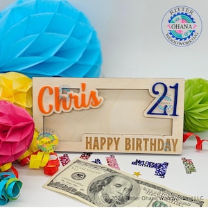 May include: A wooden picture frame with a cutout for a photo. The frame is decorated with the name "Chris" in orange letters and the number "21" in blue letters. The words "Happy Birthday" are engraved on the bottom of the frame.