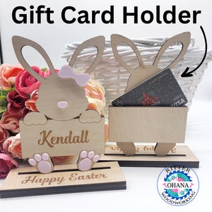 May include: Wooden gift card holder shaped like two bunnies. The bunny on the left has a pink bow and says "Kendall" and "Happy Easter". The bunny on the right has a slot for a gift card and says "A Gift For You".