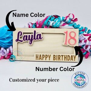 May include: A wooden sign with the words "Happy Birthday" in gold lettering. The sign is personalized with the name "Layla" in purple and the number "18" in pink.
