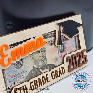 Puede incluir: Un marco de madera con un recorte de gorro de graduaci&oacute;n, un billete de d&oacute;lar y el texto "Emma 5th Grade Grad 2025". El marco est&aacute; hecho por Ritter Ohana Woodworking LLC.