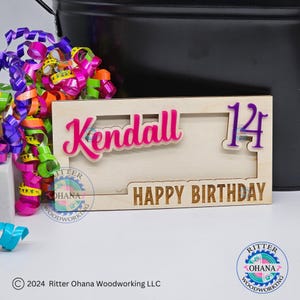 May include: A wooden birthday sign with a pink "Kendall" and a purple "14" in a cutout frame. The sign also says "Happy Birthday" in a brown font.