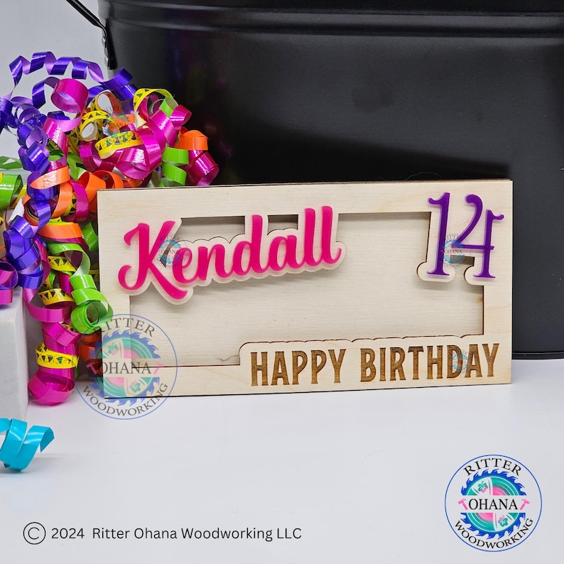 May include: A wooden birthday sign with a pink "Kendall" and a purple "14" in a cutout frame. The sign also says "Happy Birthday" in a brown font.