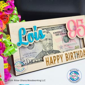 May include: A wooden frame with a cutout for a dollar bill. The frame is decorated with the name "Lois" in blue and the number "95" in pink. The text "Happy Birthday" is engraved on the frame.