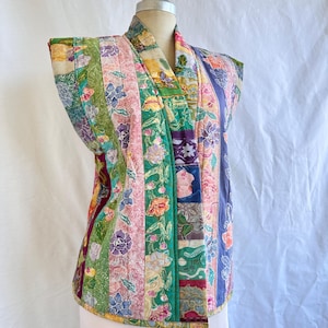 May include: A patchwork quilted vest with a variety of floral patterns in shades of pink, green, purple, and yellow. The sleeveless vest has a shawl collar and is made up of vertical panels.