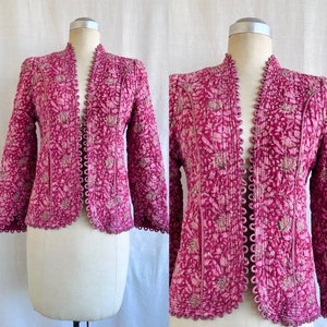 Puede incluir: Chaqueta acolchada magenta con un estampado floral. La chaqueta presenta un cuello alzado y un borde festoneado. Las mangas largas y la parte delantera abierta están adornadas con una costura decorativa. La chaqueta se muestra en un maniquí.