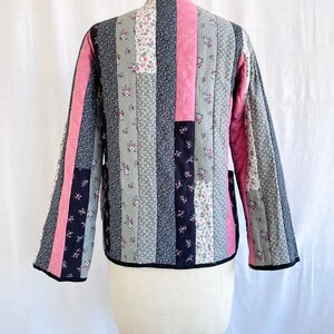 May include: A quilted jacket with a patchwork design featuring vertical stripes of pink, black and grey floral fabric. The jacket has a stand-up collar and long sleeves.
