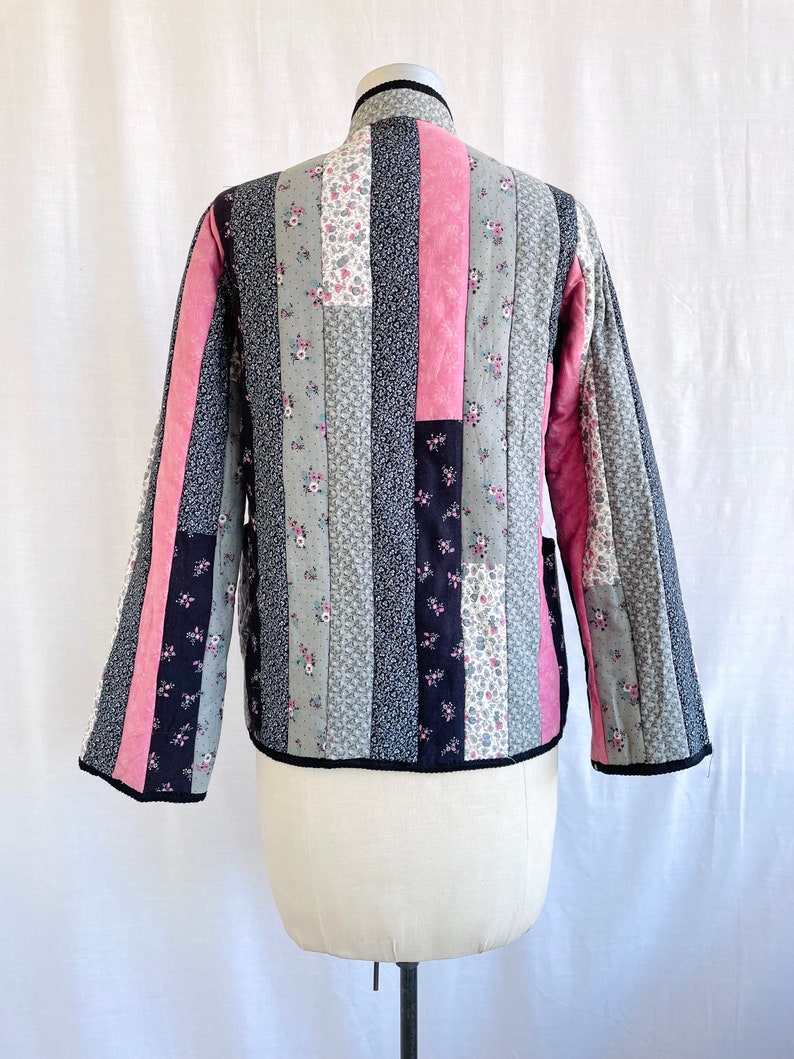 May include: A quilted jacket with a patchwork design featuring vertical stripes of pink, black and grey floral fabric. The jacket has a stand-up collar and long sleeves.