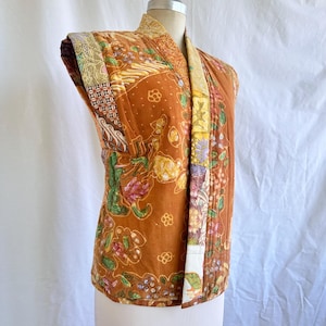 Pode incluir: Casaco sem mangas acolchoado em tons de castanho, laranja e amarelo, com um design de patchwork com padrões florais e geométricos. O casaco é exibido num manequim, mostrando a sua frente aberta e combinações de tecidos únicas.