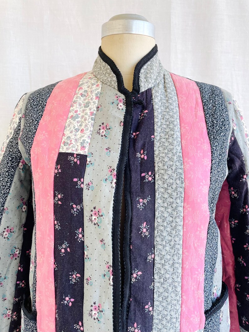 May include: A quilted jacket with a mandarin collar and a button fastening. The jacket is made from patchwork fabric in shades of pink, black and grey with floral patterns.