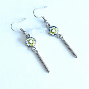 May include: A pair of drop earrings featuring silver-toned metal hooks and a dangling cylindrical bar. Each earring includes a circular charm with a white, yellow, and blue floral pattern, accompanied by two small clear plastic earring backs.