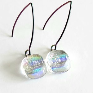 May include: A pair of iridescent glass earrings with a square shape and a subtle, textured design. The earrings hang from long, curved, dark metal ear wires. The glass has a shimmering, multi-coloured effect.