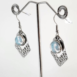 May include: A pair of silver-coloured dangle earrings. Each earring has a round, light blue glittery centre set within a hammered diamond-shaped frame. The earrings hang from a simple hook.