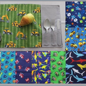 May include: A green and grey fabric roll-up featuring a construction vehicle design, holding a pear and cutlery. Additional fabric swatches show space, construction, dinosaur, and shark patterns. The roll-up is designed to hold utensils.
