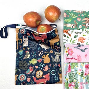 May include: A blue reusable snack bag with a zipper closure and a navy blue handle. The bag is printed with a colourful pattern of animals, flowers, and leaves. Three red apples are visible in the background.