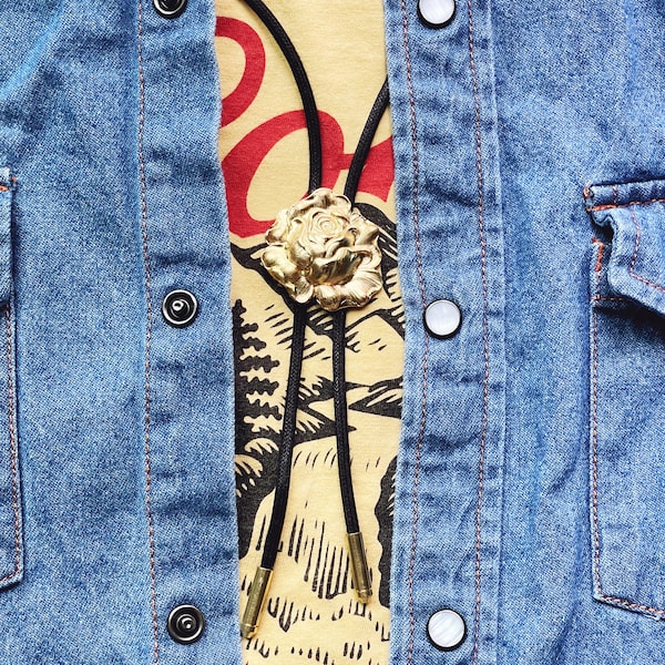 Könnte beinhalten: Ein schwarzes Bolo-Band mit einem goldenen Rosenanhänger. Das Band wird über einem gelben T-Shirt mit einem schwarz-weißen Design und einer blauen Jeansjacke getragen.