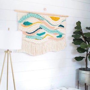 May include: A white woven wall hanging with a wood dowel at the top. The hanging features a wavy pattern in shades of blue, yellow, and pink. The hanging has a fringe at the bottom.